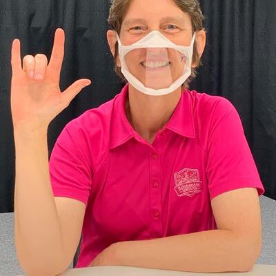 Cheryl Gault, Part-time Office Coordinator, wears a clear face mask and signs "I Love You" in ASL